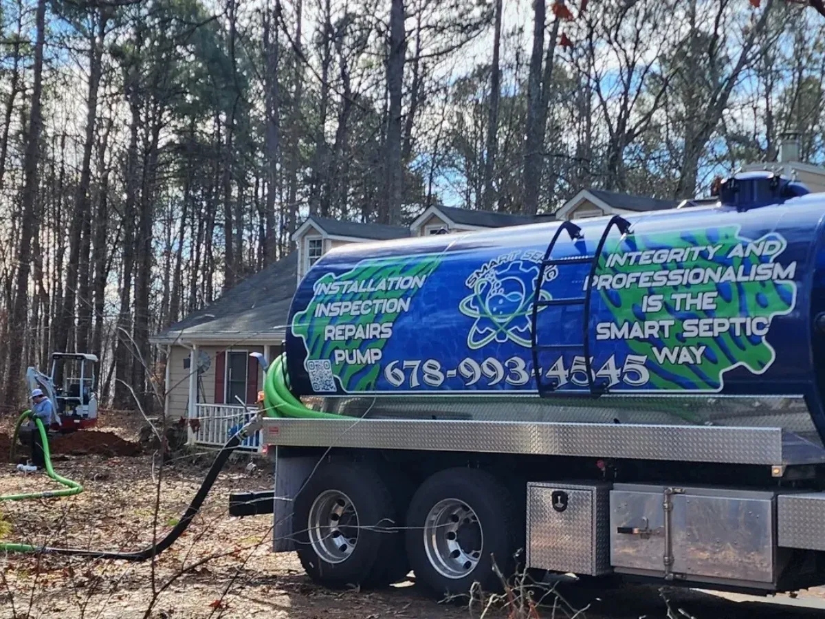 A septic service truck with hoses extended is parked in front of a house surrounded by trees. A worker operates equipment in the yard, ensuring proper flow and considering the use of septic additives for system maintenance.