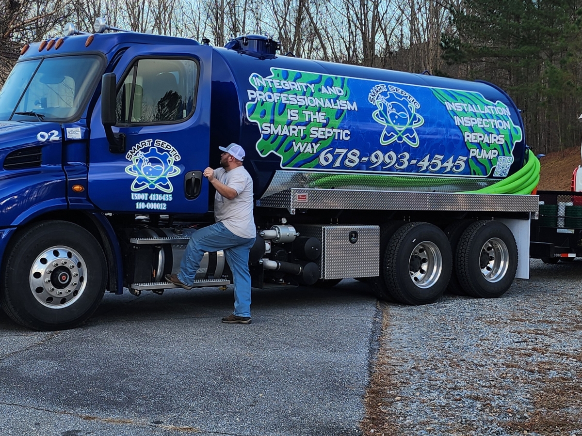 A man in a white shirt and cap steps onto a blue septic service truck with company information, services, and details about septic additives written on the side.