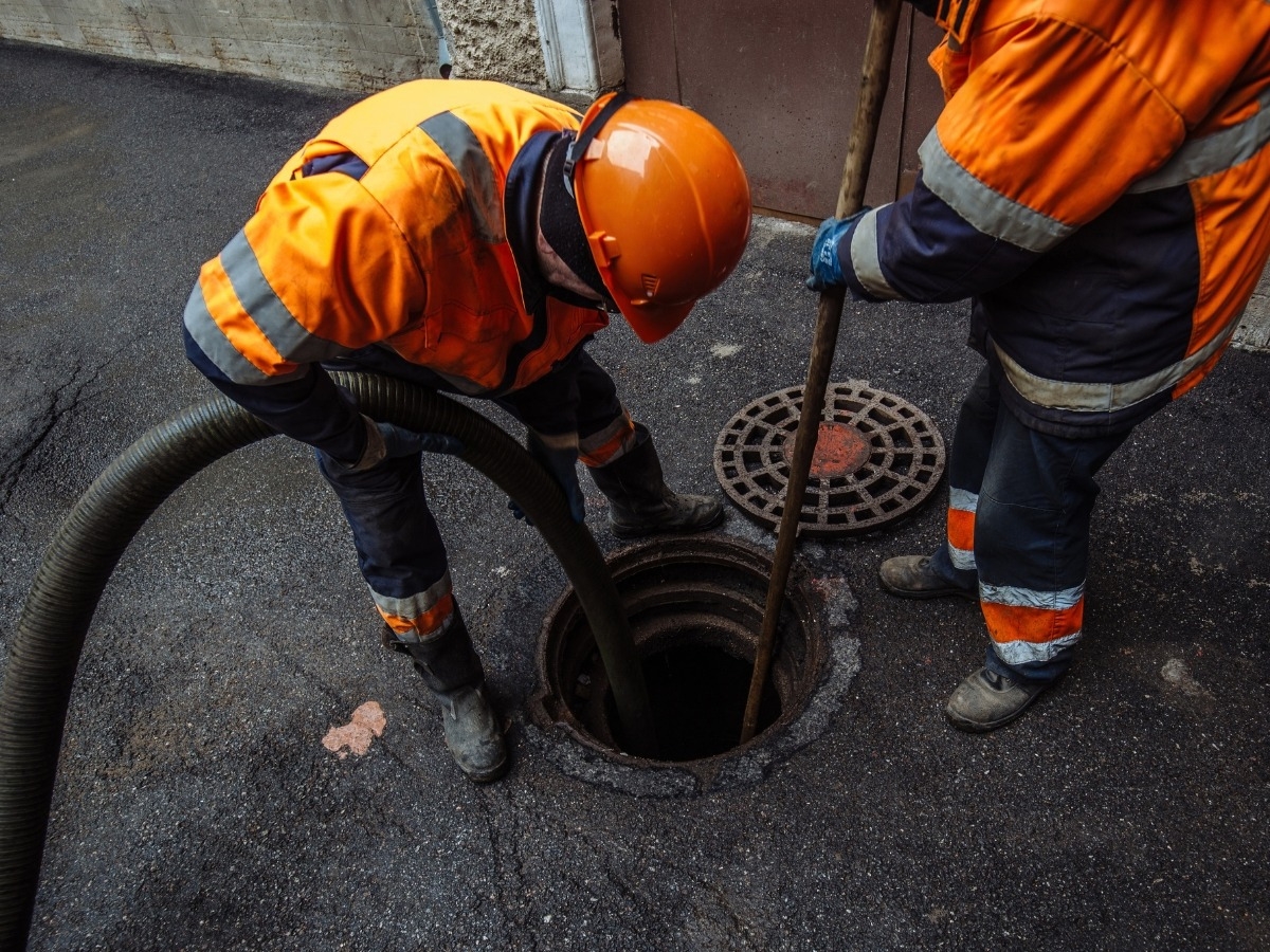 Two workers in orange safety gear clean a sewer with a hose and tool, standing by an open manhole on asphalt. Septic additives may be used alongside such maintenance to enhance sewer system performance.