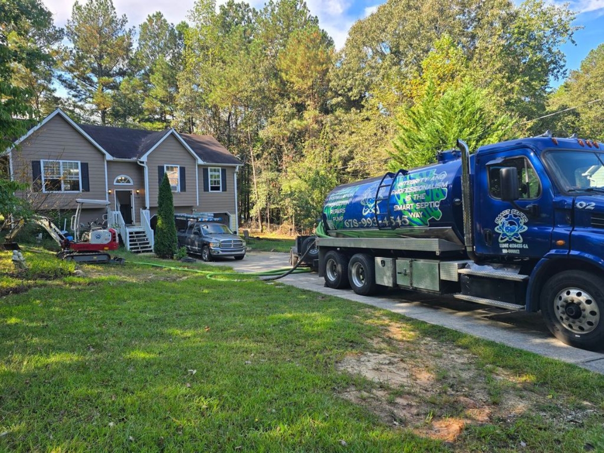 A septic service truck, essential for emergency prep, is parked in the driveway of a suburban house with equipment set up on the lawn. Trees and grass surround the property on a sunny day.