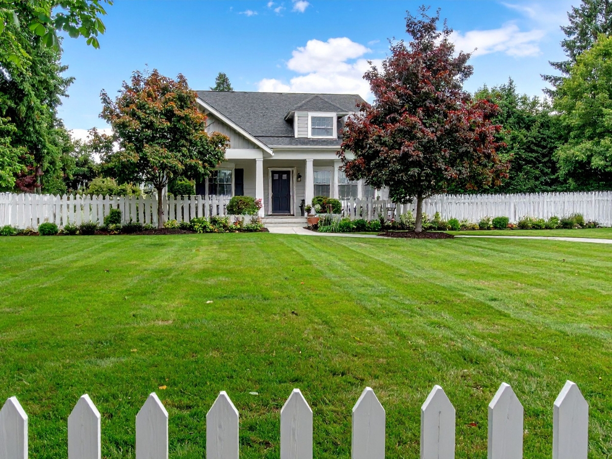 Landscaping Near Septic Systems: What to Plant & Avoid 2 A single-story house with a front porch, surrounded by a white picket fence, manicured lawn, well-designed landscaping, green trees, and a partly cloudy sky.