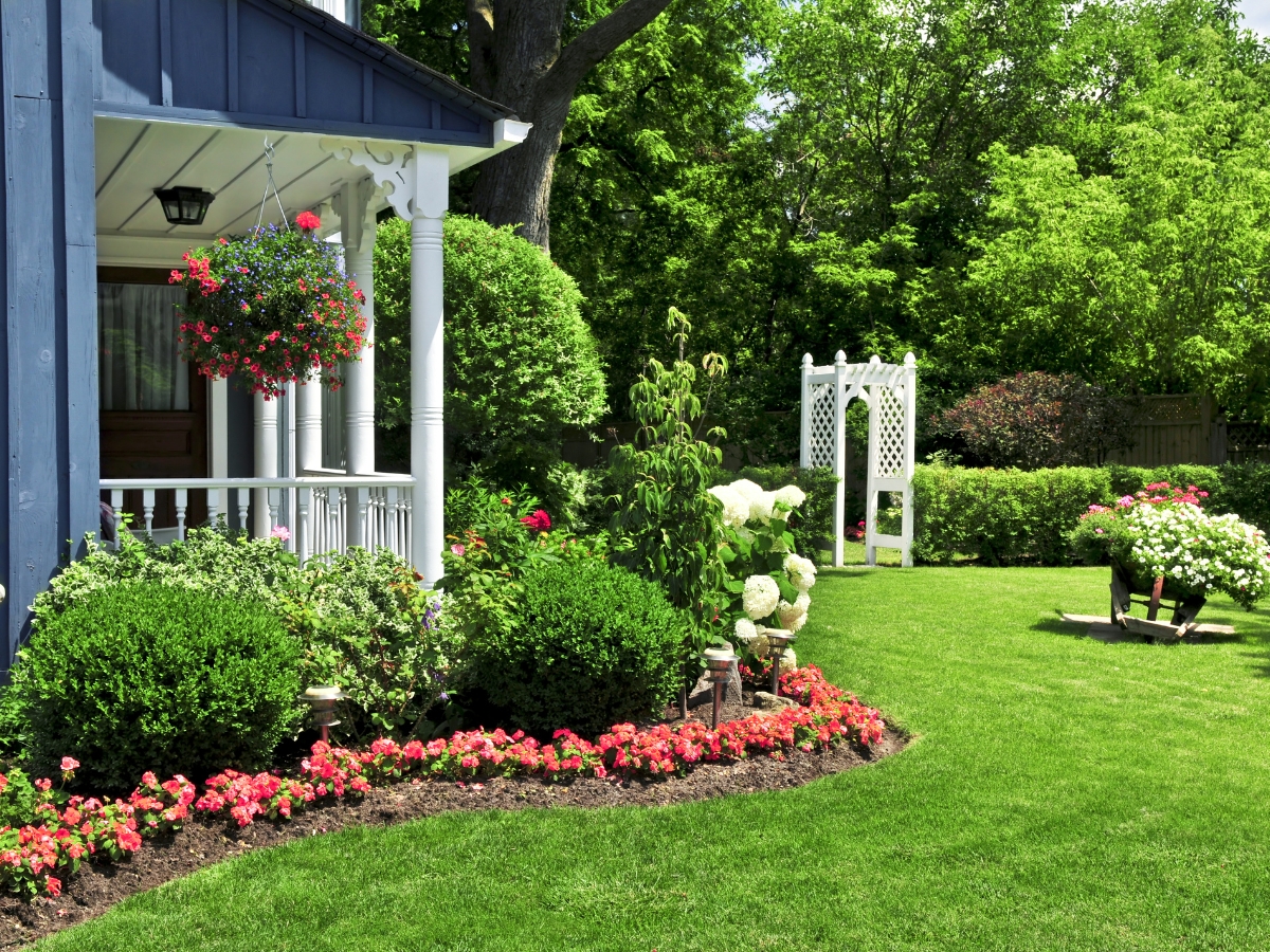 Landscaping Near Septic Systems: What to Plant & Avoid 3 A well-kept garden with pink flowers, trimmed bushes, expert landscaping, a blue house porch, hanging basket, green lawn, and a white garden arbor in the background.