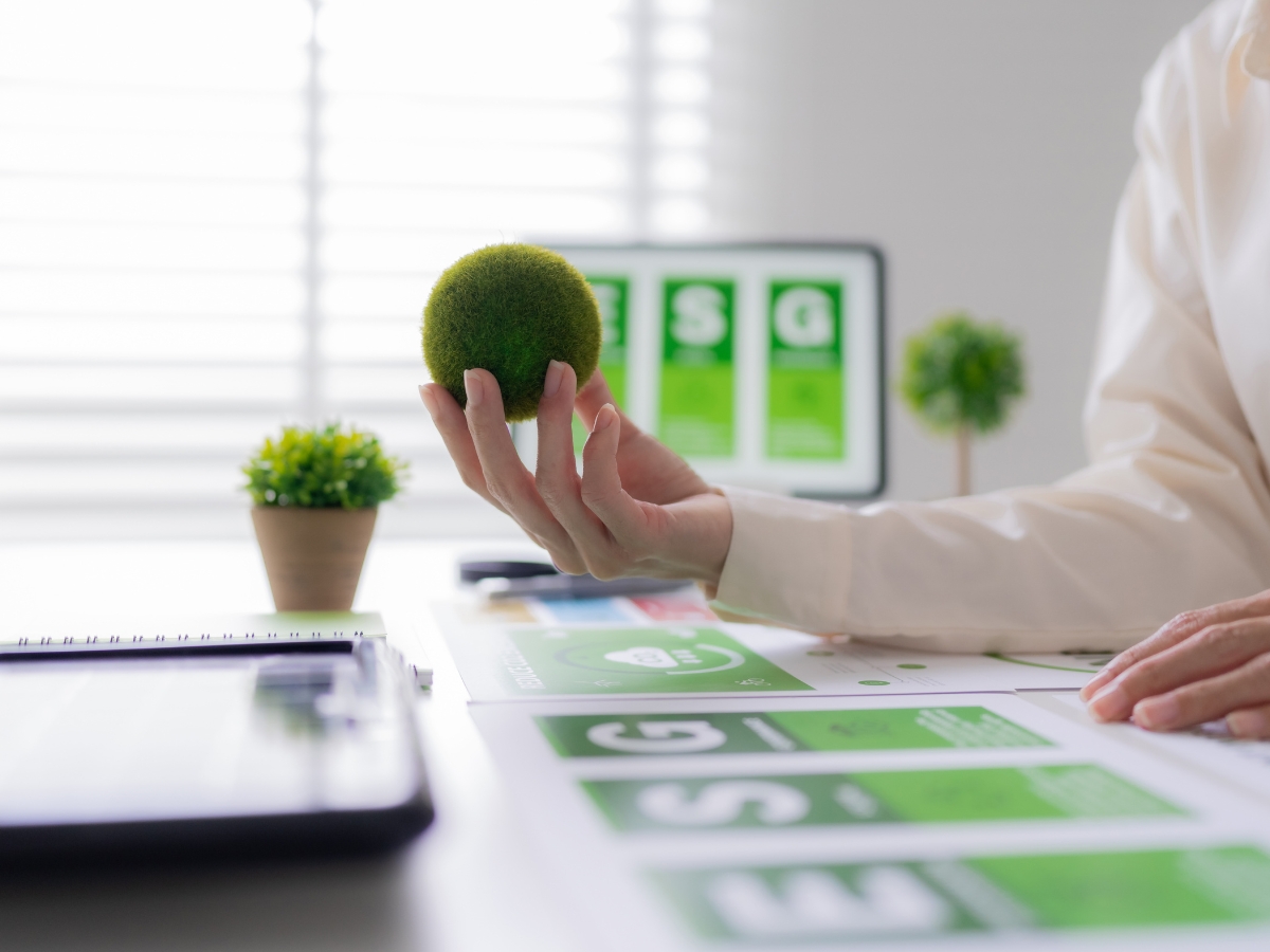 Person holding a small green moss ball at a desk with ESG documents, a laptop, and a computer screen displaying ESG graphics and New Technologies in an office setting.