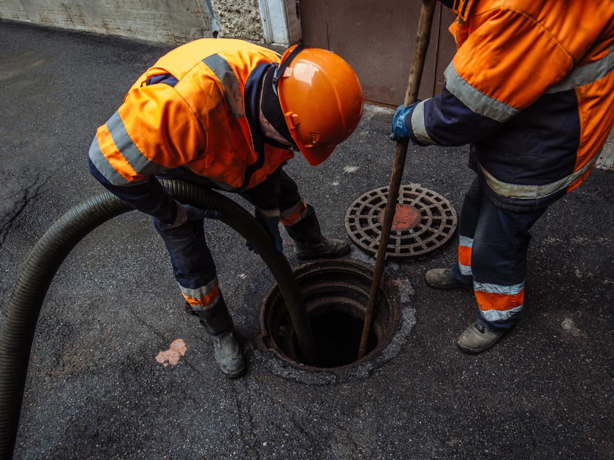 Two workers in orange safety gear clean or inspect a sewer drain, possibly as part of real estate inspections, using a large hose and a pole near an open manhole on an asphalt surface.