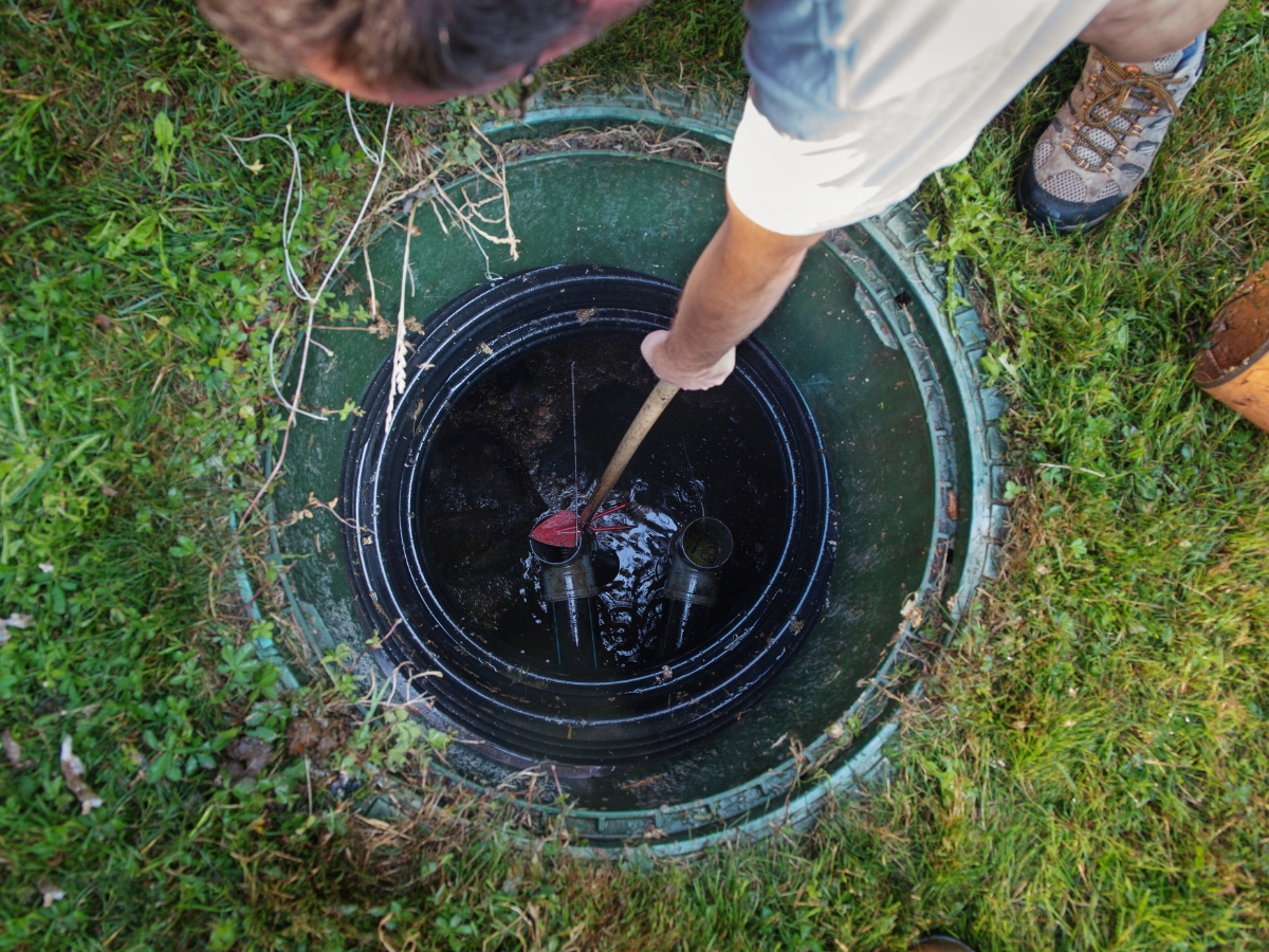 A person wearing boots and shorts inspects the inside of a green outdoor tank or well, viewed from above, using a wooden stick—demonstrating proper septic system care.