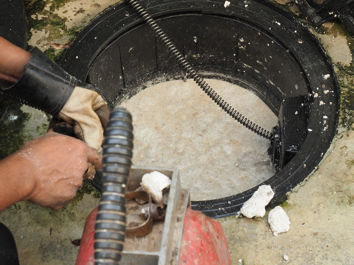 A person uses a plumbing snake to clear a blocked drain, with debris and white residue around the opening—an important step in septic system care to prevent further clogs and maintain efficient drainage.