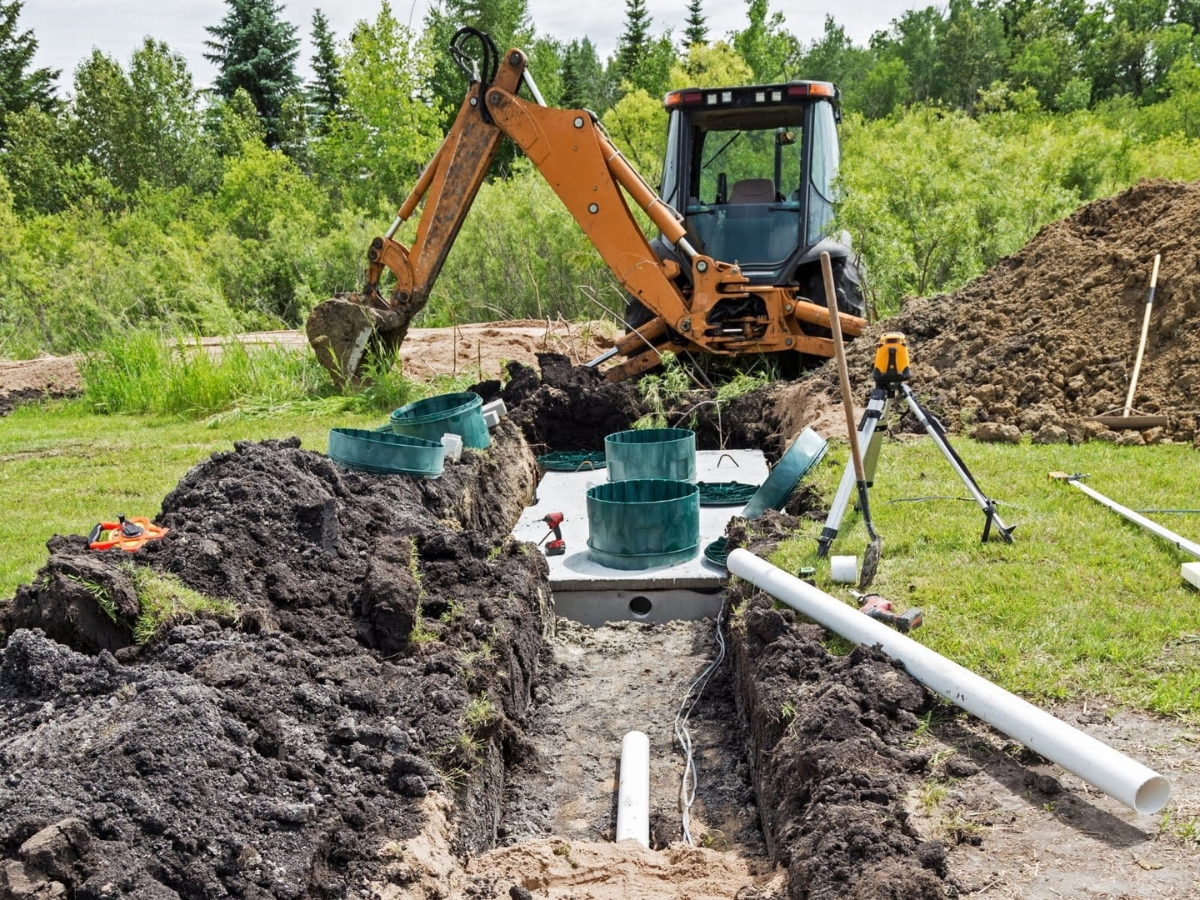 A backhoe excavates soil at a construction site, demonstrating proper septic system care with exposed pipes and septic components on a grassy area surrounded by trees.